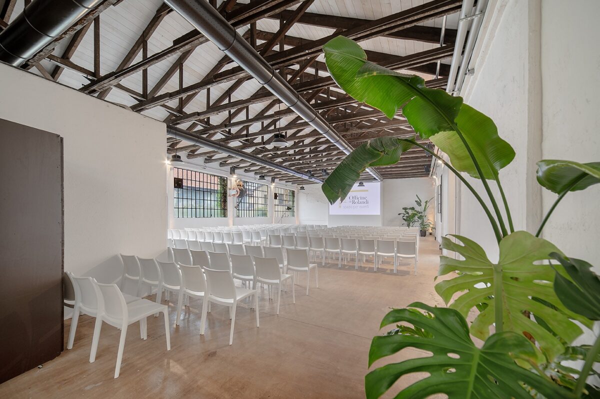 sala principale allestita a platea con sedie bianche e schermo acceso, grandi vetrate illuminano la sala